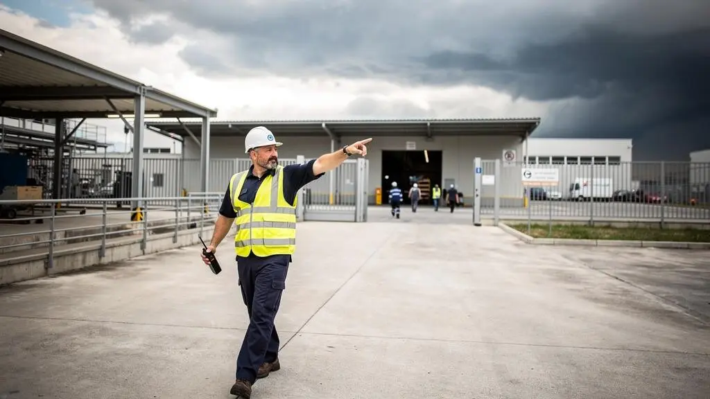 Supervisore della sicurezza che dirige l'evacuazione preventiva di lavoratori da area esterna industriale durante allerta fulmini